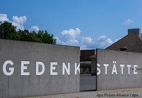Mit der WebApp „Dingen auf der Spur“ Gedenkstättenbesuche vorbereiten: Bild der Gedenkstätte und des Museum Sachsenhausen. Im Vordergrund ist eine unterbrochene Mauer aus Beton zu sehen, die aus zwei Teilen besteht. Auf den Teilstücken ist in großen weißen Buchstaben das Wort "Gedenkstätte" zu lesen. Hinter der Mauer sind Häuser als Teile der Gedenkstätte zu erkennen.