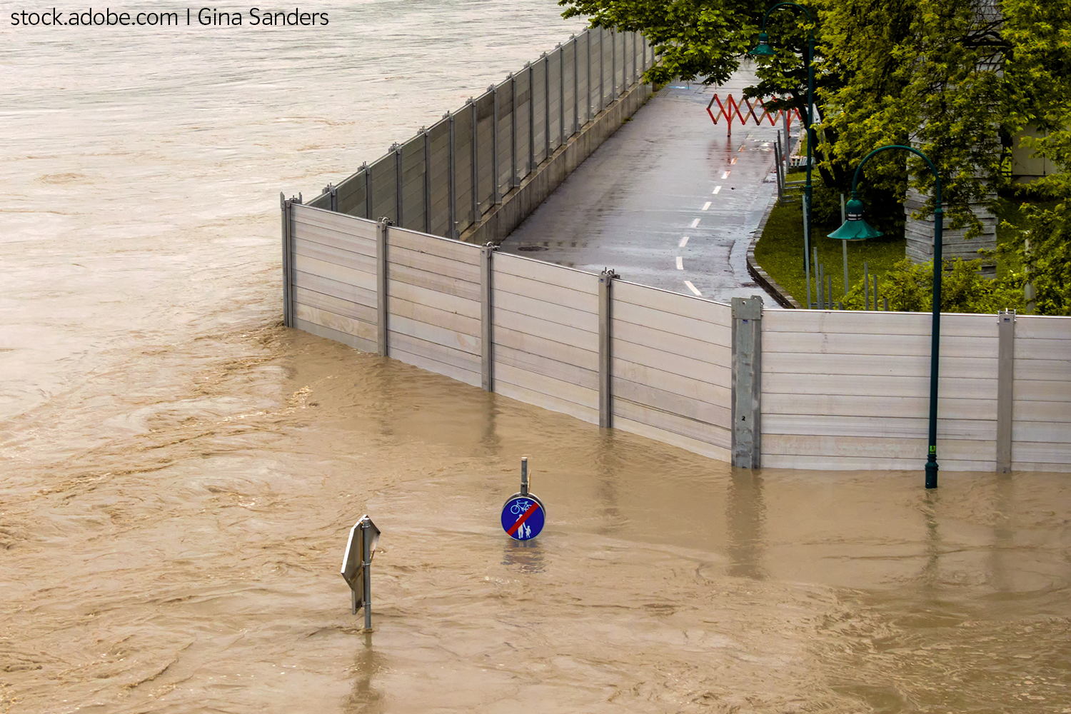 Das Bild zeigt einen Straßenabschnitt, der von einer mobilen Hochwasserschutzmauer vor Hochwasser geschützt ist. Hinter der Mauer ist es trocken bzw. kein Wasser, während vor der Mauer alles von braun-trübem Wasser überflutet ist. Das Wasser steht so hoch, dass es die untere Hälfte der Schutzmauer bedeckt und dass gerade noch die Verkehrszeichen aus den Fluten herausschauen. Alles andere ist überflutet.