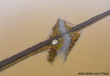 Der Kreuzungspunkt einer Straße und einer Eisenbahntrasse ist fast völlig überflutet. Nur ein kleiner Bereich mit der Kreuzung, einem Bahnhäuschen und etwas Wiese ist noch nicht vom Wasser bedeckt. Der Ausschnitt des Bildes zeigt nur diesen Knotenpunkt, rundherum umgeben von trübem Wasser.