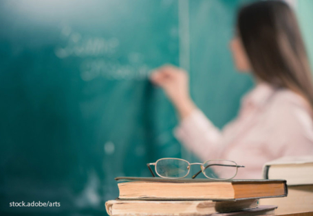 Brille liegt auf Bücherstapel, Lehrerin schreibt auf Tafel im Hintergrund.